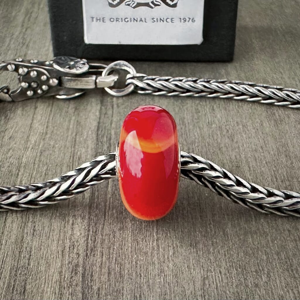 Available at Suzie Q Studio dot com, Trollbeads retired SCARLET ARMADILLO, glass bead, in vibrant red and orange, shown on a silver chain with a clasp.