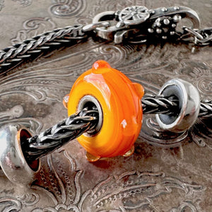 Trollbeads STRIPED BUD is a yellow-orange glass bead with a mix of whimsical stripes and clear dots, shown on a Trollbeads silver bracelet.